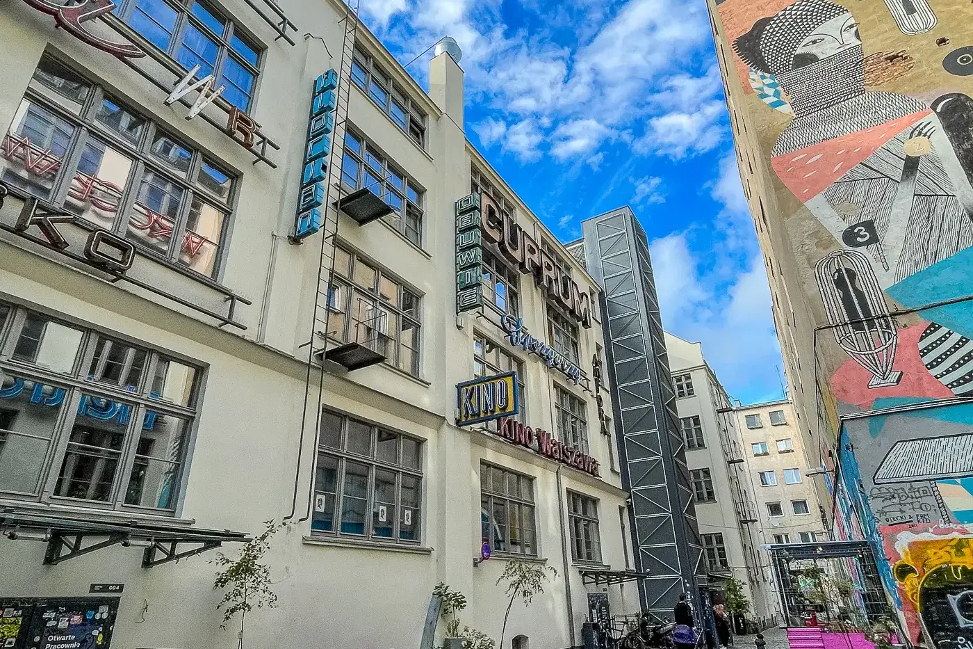 A narrow cobbled alleyway in Wrocław, flanked by a building with neon signs and another with a large abstract mural. Outdo...