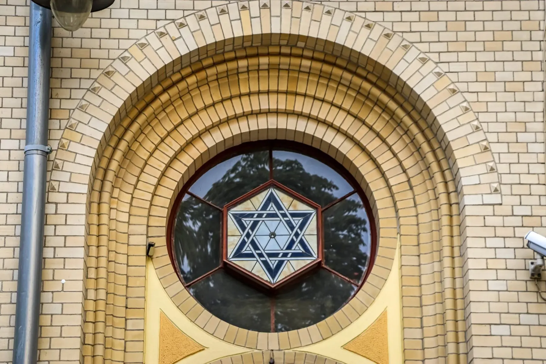 Close-up of a round window featuring a Star of David symbol, set within an arched light brick facade. A metal gutter is on...