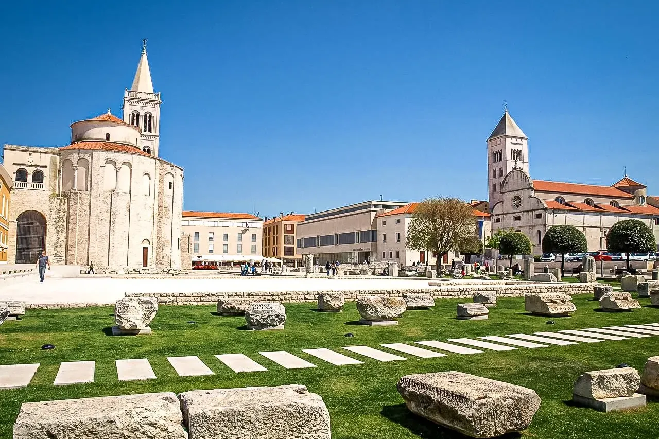 View of Zadar's Roman Forum ruins with grass, paving stones, and stone blocks. In the background are the Church of St. Don...