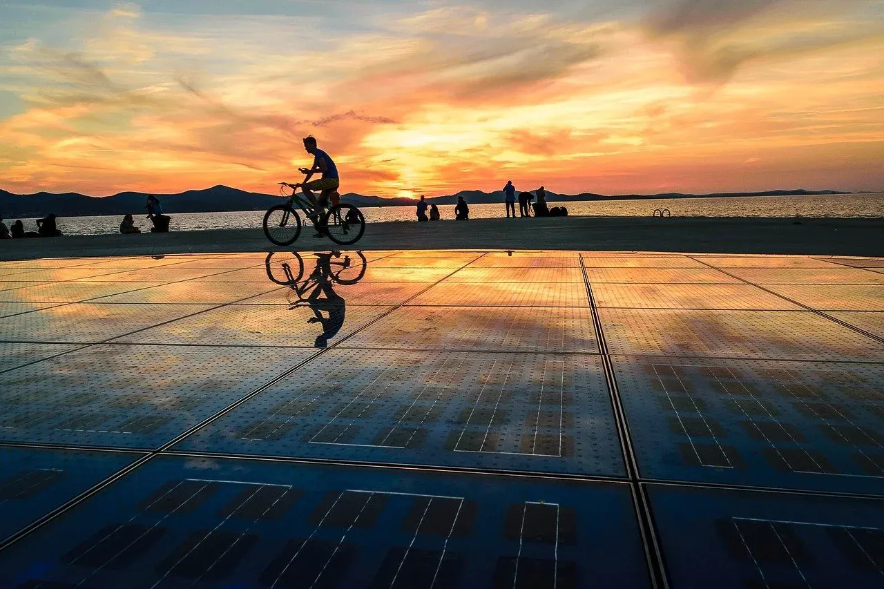Person on bicycle silhouetted against a colorful sunset over the sea, reflecting on a large solar panel art installation.