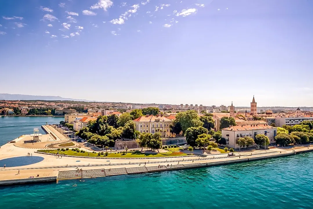 Aerial view of Zadar waterfront with the Sea Organ, old town buildings, clear blue sea, and distant mountains under a sunn...