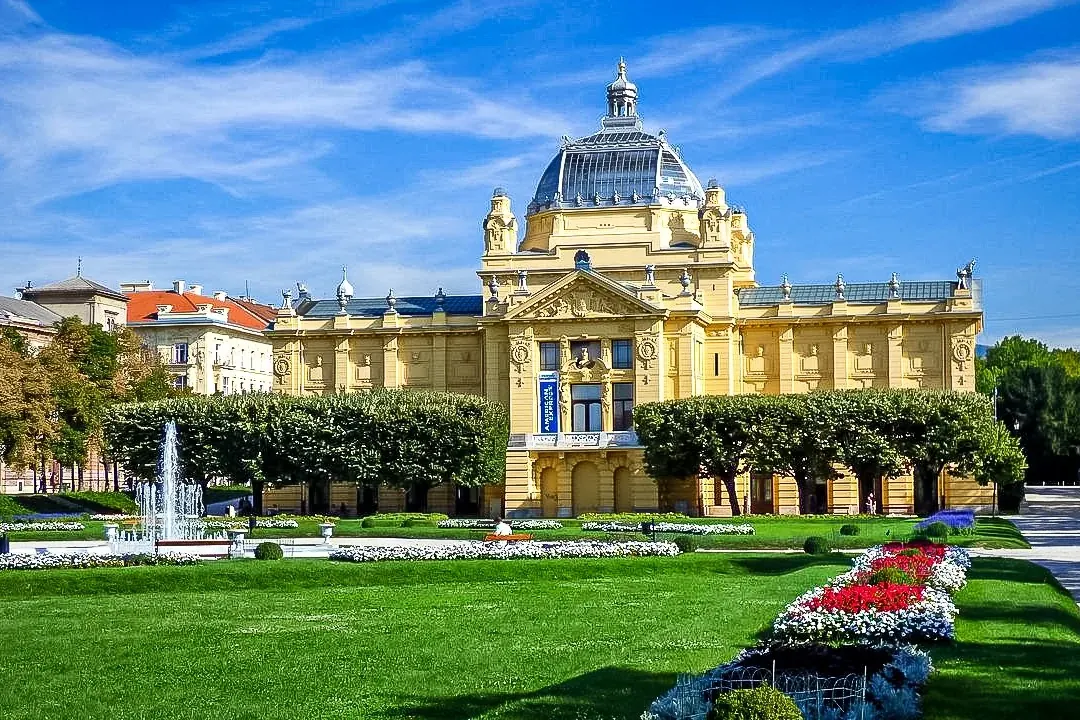 Yellow Neo-Baroque Art Pavilion with a glass dome, a fountain in a green park with flower beds, and a distant cathedral sp...
