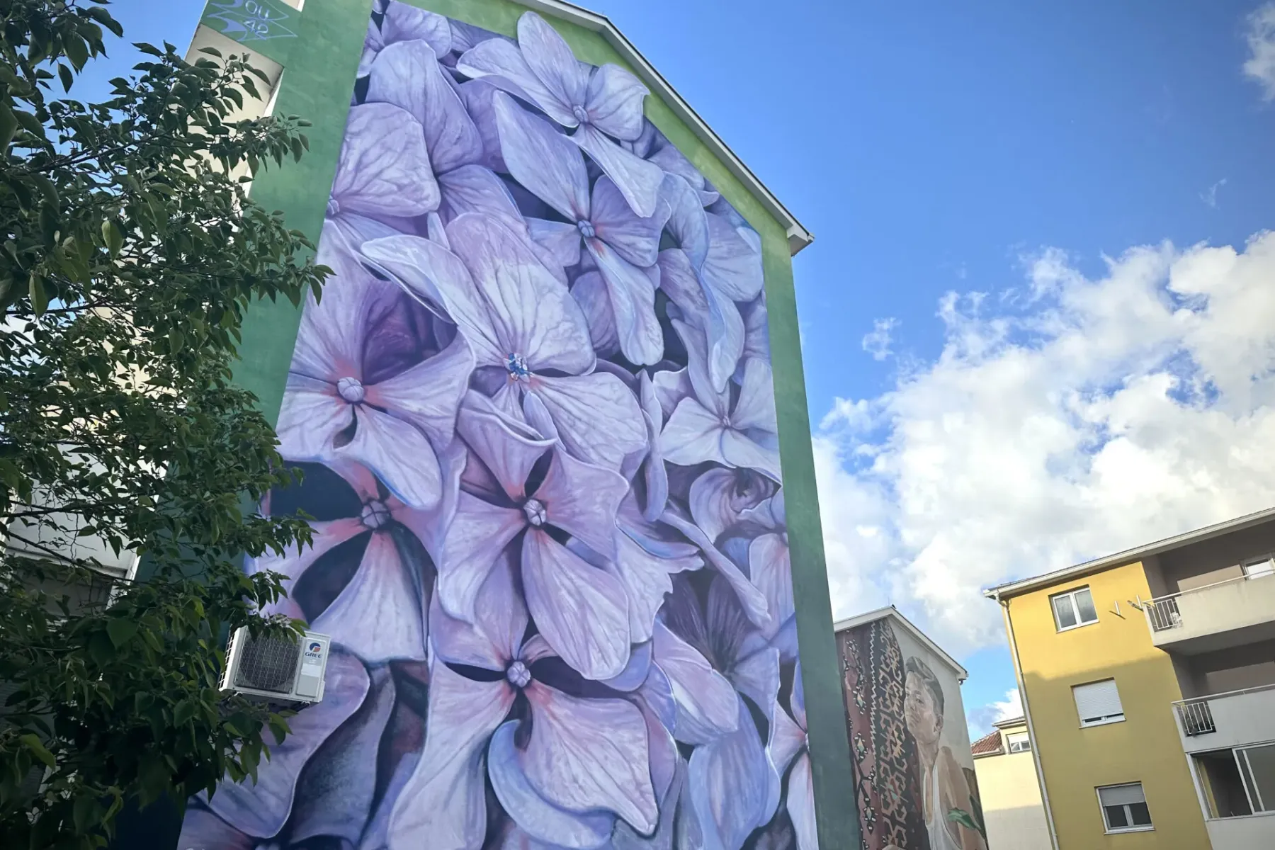 Large mural of purple hydrangeas on a green building facade. A tree branch and air conditioning unit are visible on the left.