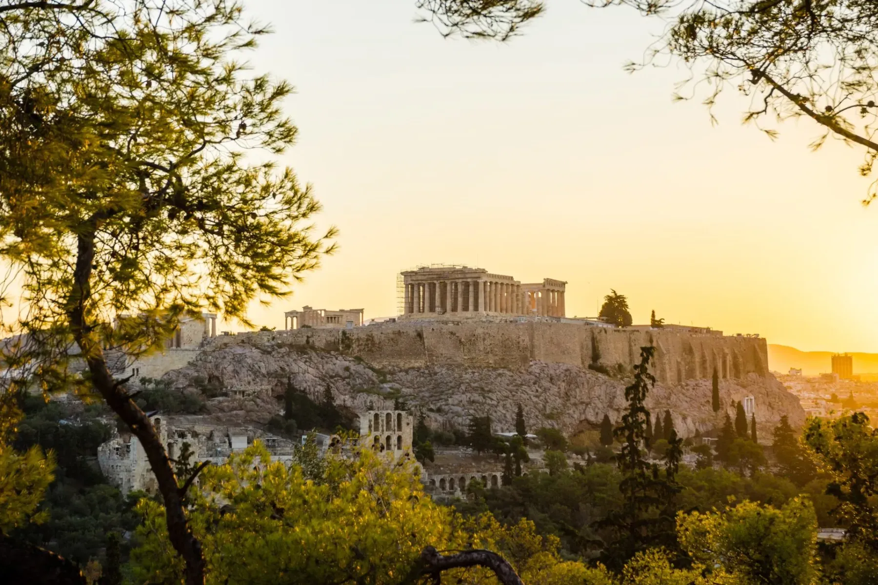 The Acropolis of Athens with the Parthenon and scaffolding, bathed in warm sunset light, seen through green trees.