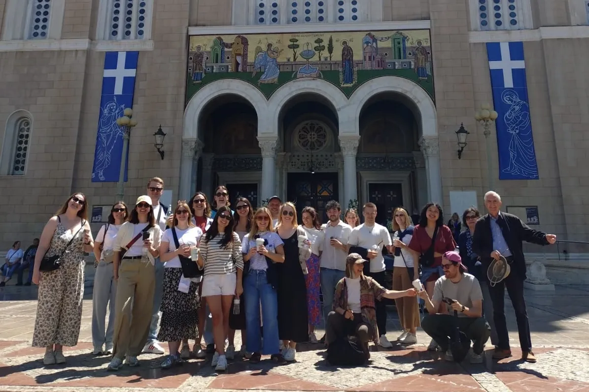 A tour group smiles for a photo in front of the Metropolitan Cathedral of Athens on a sunny day.
