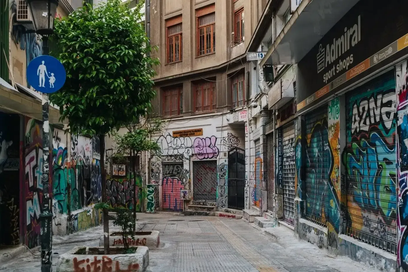 Narrow urban alley with buildings and shop shutters covered in colorful graffiti. Small trees in planters and a pedestrian...