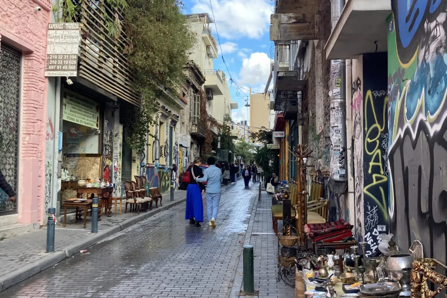Paved street in Athens with buildings covered in graffiti and a flea market stall on the right. Two people walk away.