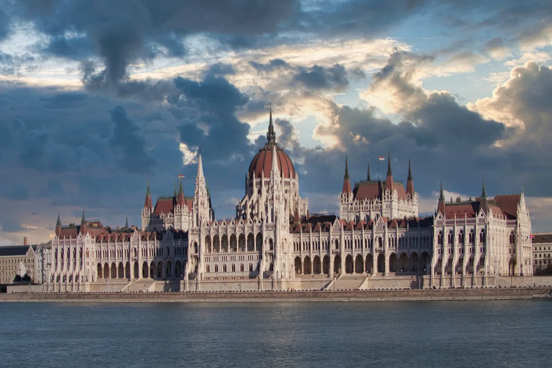 The Hungarian Parliament Building viewed from across the Danube River under a dramatic, cloudy sky.