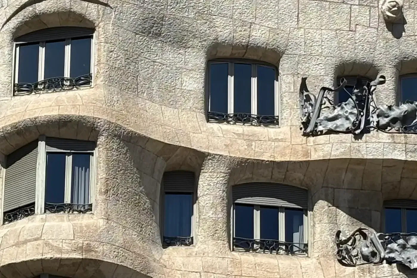 Close-up of Casa Milà's wavy stone facade with dark windows, grey roller blinds, and wrought-iron balconies under a blue sky.