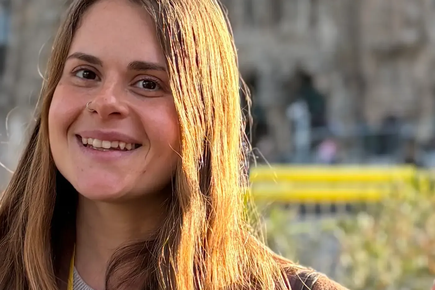 Smiling woman with long brown hair, brown coat, and yellow 'Catalonia Tourism Guide' lanyard. Blurry building and palms in...