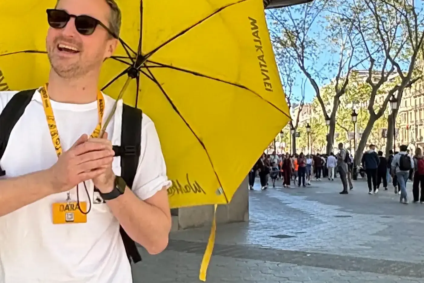 A smiling male tour guide holding a yellow Walkative umbrella on a sunny street in Barcelona. Ornate modernist building in...