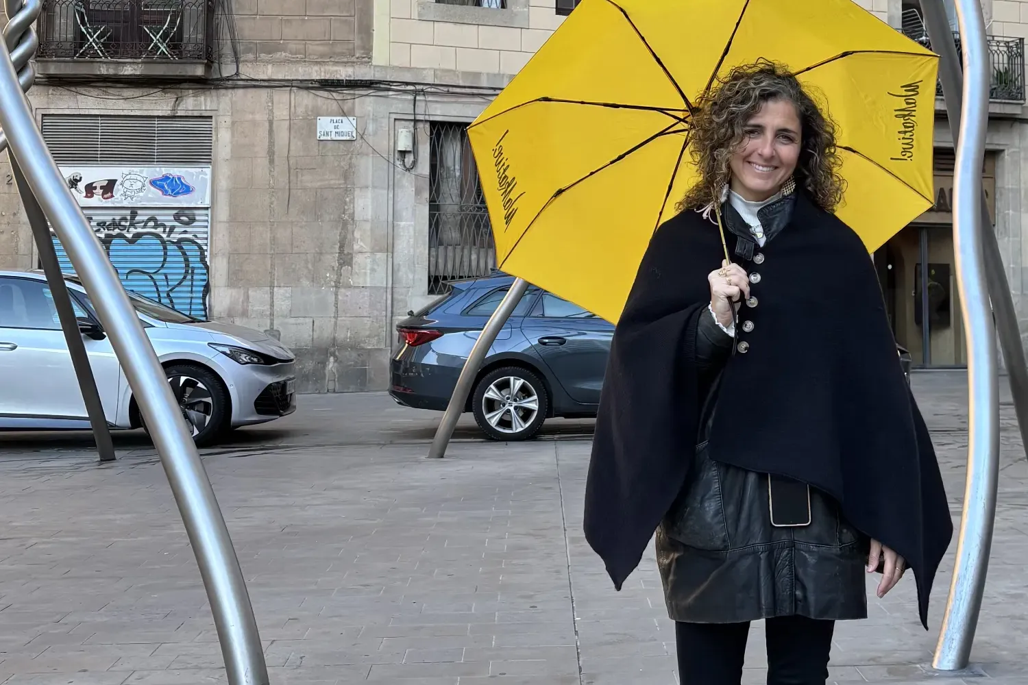 Smiling woman with curly hair holds a yellow Walkative umbrella in Plaça de Sant Miquel, Barcelona. Buildings and cars beh...