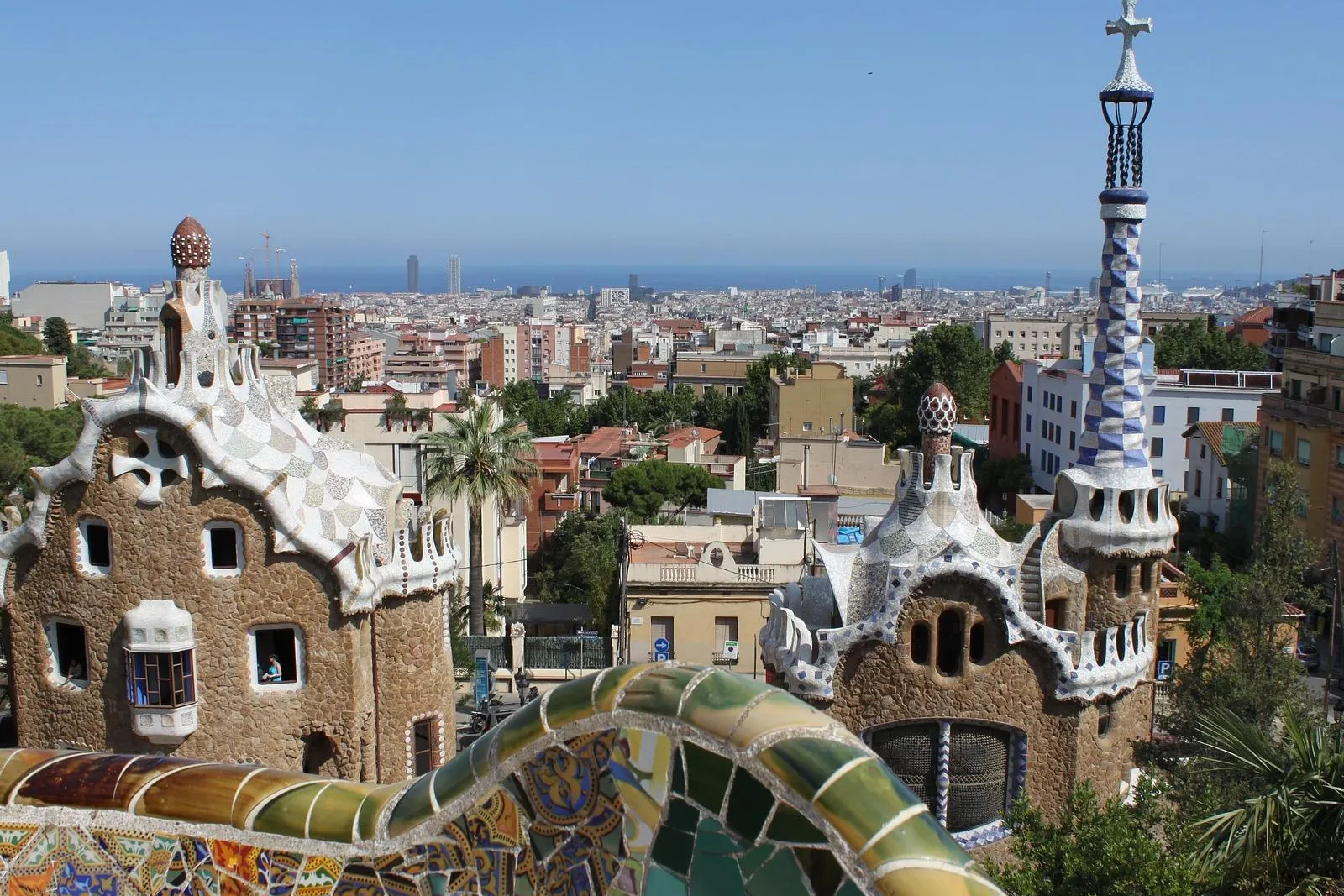 View from Park Güell showing two Gaudi-designed mosaic-roofed buildings, a colorful tiled wall, and Barcelona cityscape wi...