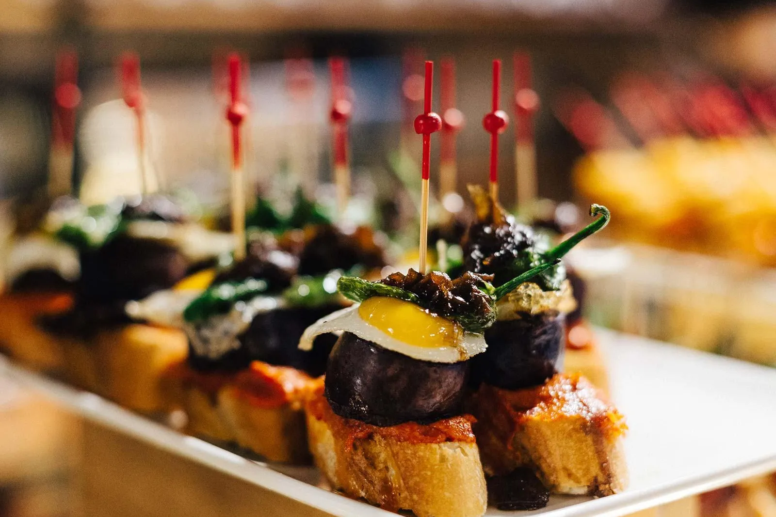 Close-up of a plate of pintxos, small open-faced sandwiches with various toppings, held by red toothpicks. A dark sausage ...