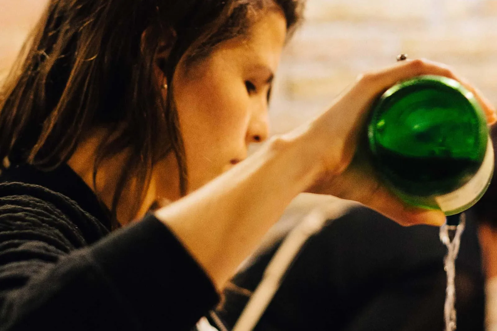 Person pouring sidra from a green bottle into a glass, generating foam. Blurred stone wall in background.