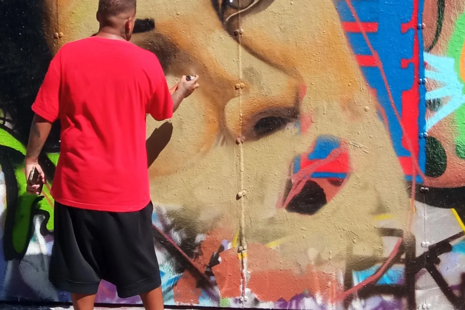 A person in a red t-shirt spray painting a large mural of a woman's face on a wall covered in graffiti. Spray cans on the ...