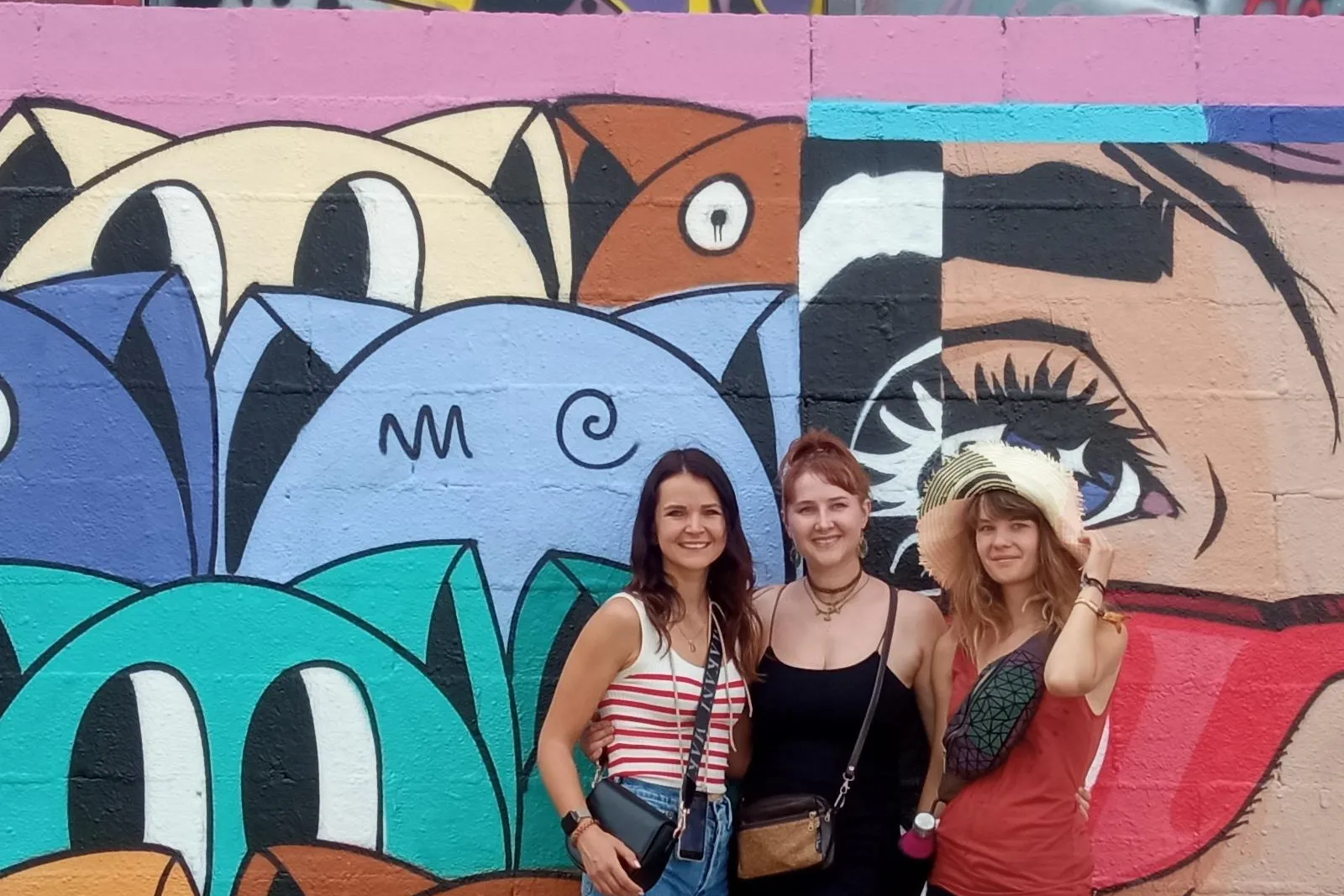 Three women smiling in front of a colorful wall covered in abstract graffiti and a large mural with stylized faces.