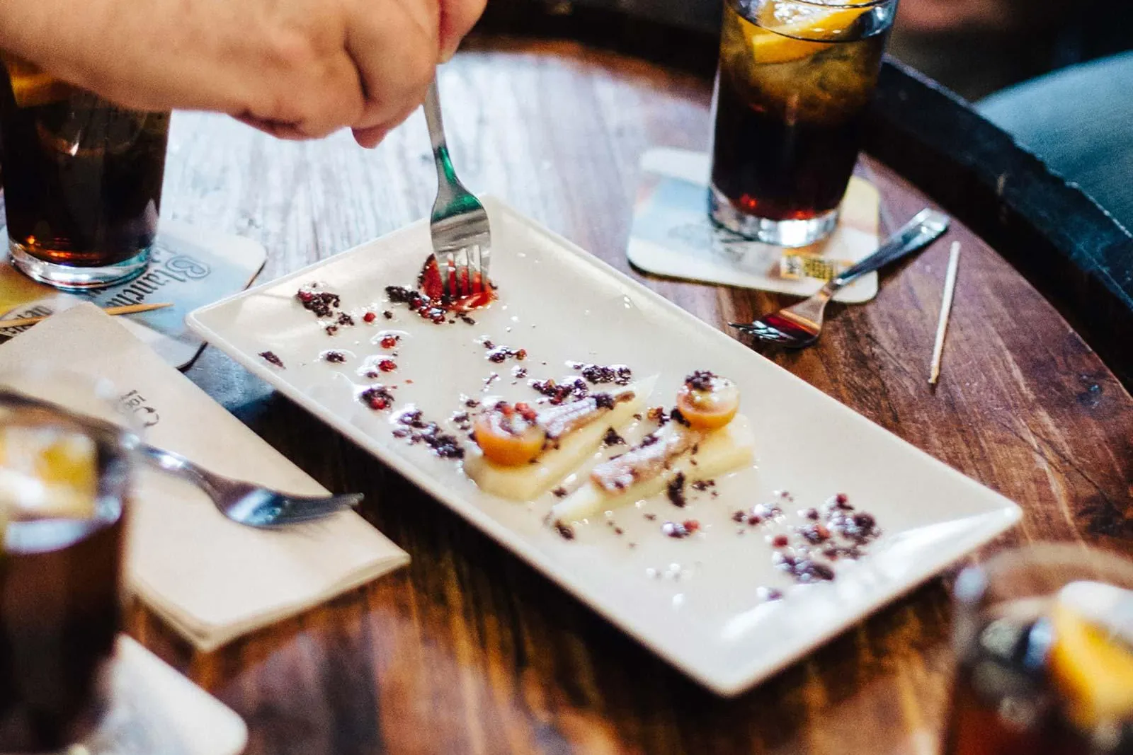 People's hands around a wooden table with tapas plates and drinks. One person uses a fork to eat a dish.
