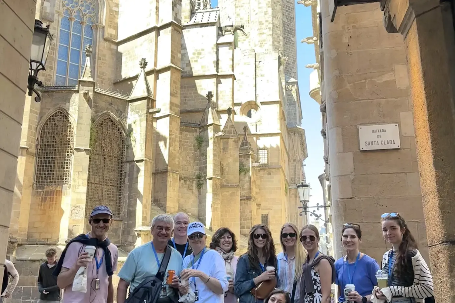 A diverse group of people stands on a narrow stone street with Barcelona Cathedral in the background under a blue sky.