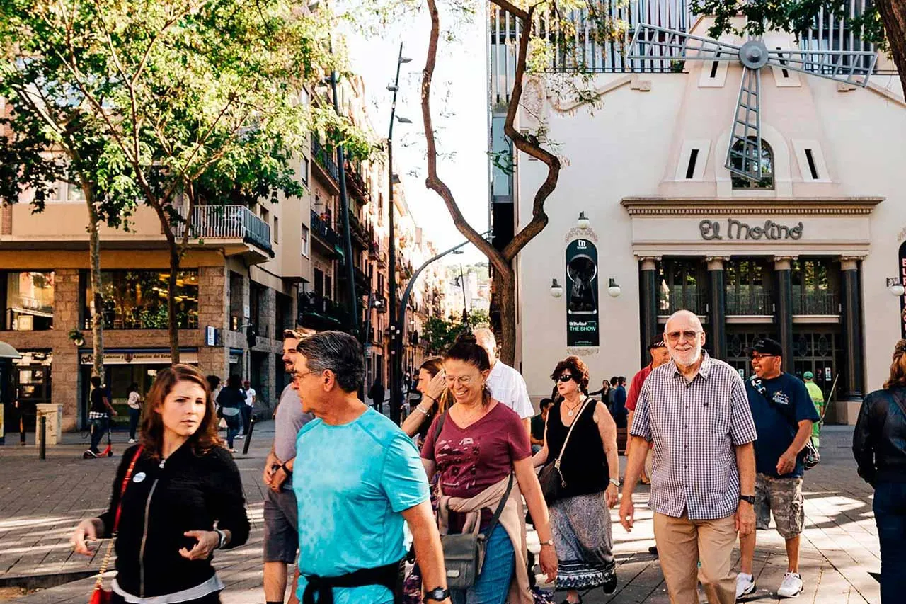 Group of people walking on a sunny street past El Molino theatre with a windmill on its facade in Barcelona.