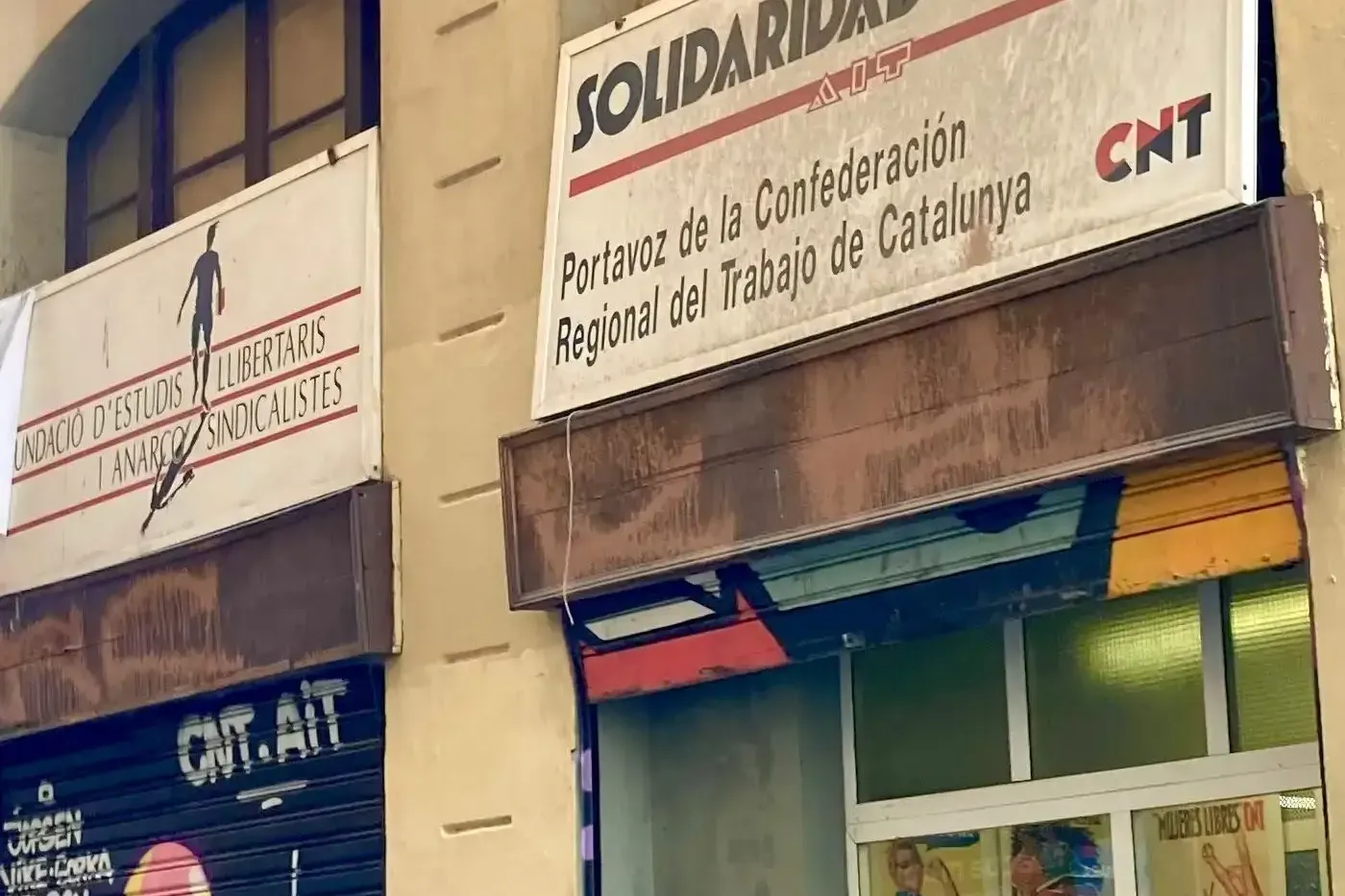 A group of people on a street tour looking up at a building with "Solidaridad Obrera" and "CNT" signs, and graffiti on a s...