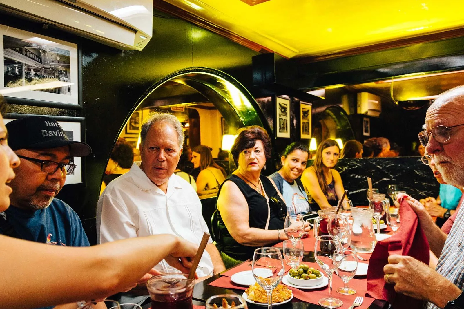 A group of people sitting at a restaurant table with red tablecloths, sharing tapas dishes, olives, and a pitcher of red s...