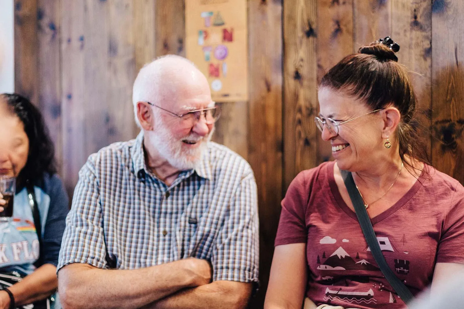 Two smiling travelers, an older man and a woman, talking in a casual setting with a wooden wall and a tapas poster.