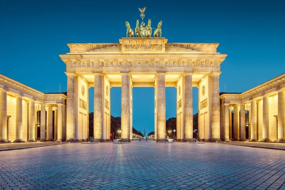 The Brandenburg Gate in Berlin, illuminated at dusk, featuring the Quadriga sculpture on top and a wide, empty cobblestone...