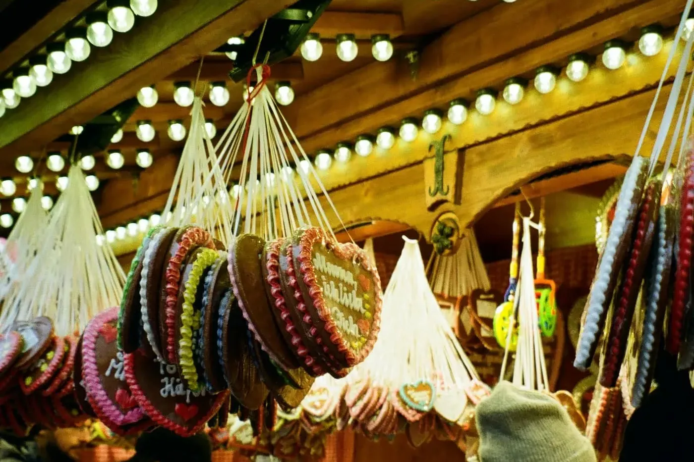 Hanging gingerbread heart cookies with icing decorations and various sweets at a festive market stall, with people browsing.