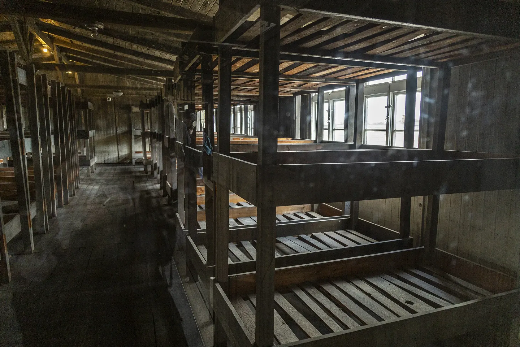 Interior of a wooden barrack with multi-tiered bunk beds, long rows extending into the dimly lit space. Windows are visibl...