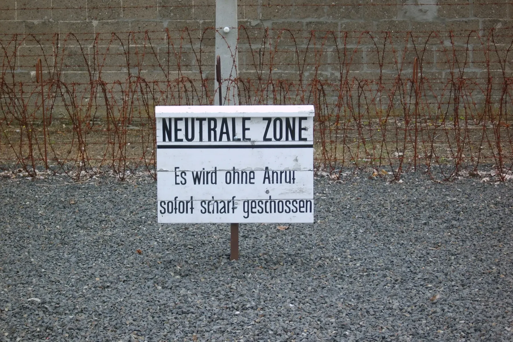 White sign with "NEUTRALE ZONE" and warning text in German, in front of barbed wire fence and concrete wall on gravel ground.