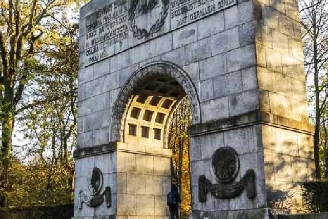 Large grey stone archway monument with carved inscriptions and reliefs, a person walks through it in a park setting.