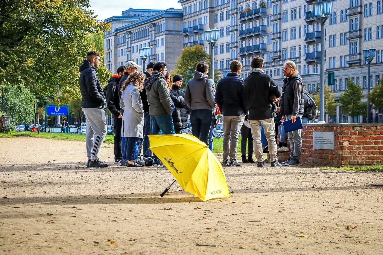 A group of people listening to a guide near apartment buildings in Berlin. A yellow Walkative umbrella rests on the ground.