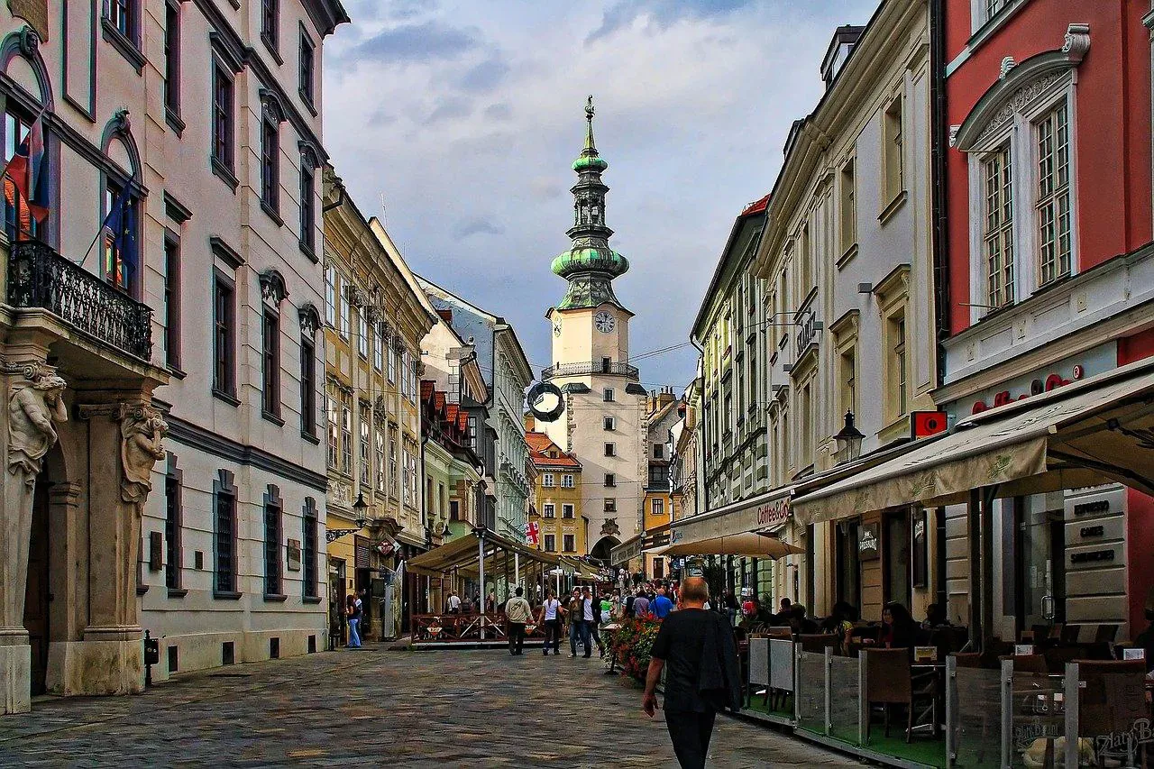 Historic street in Bratislava leading towards Michael's Gate, flanked by old buildings and outdoor cafes with people walking.