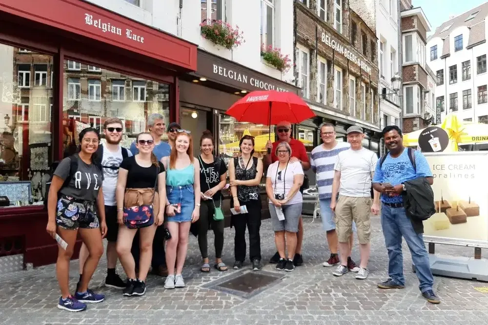 A diverse group of tourists stands smiling on a cobblestone street in front of Belgian Lace and Chocolate shops.