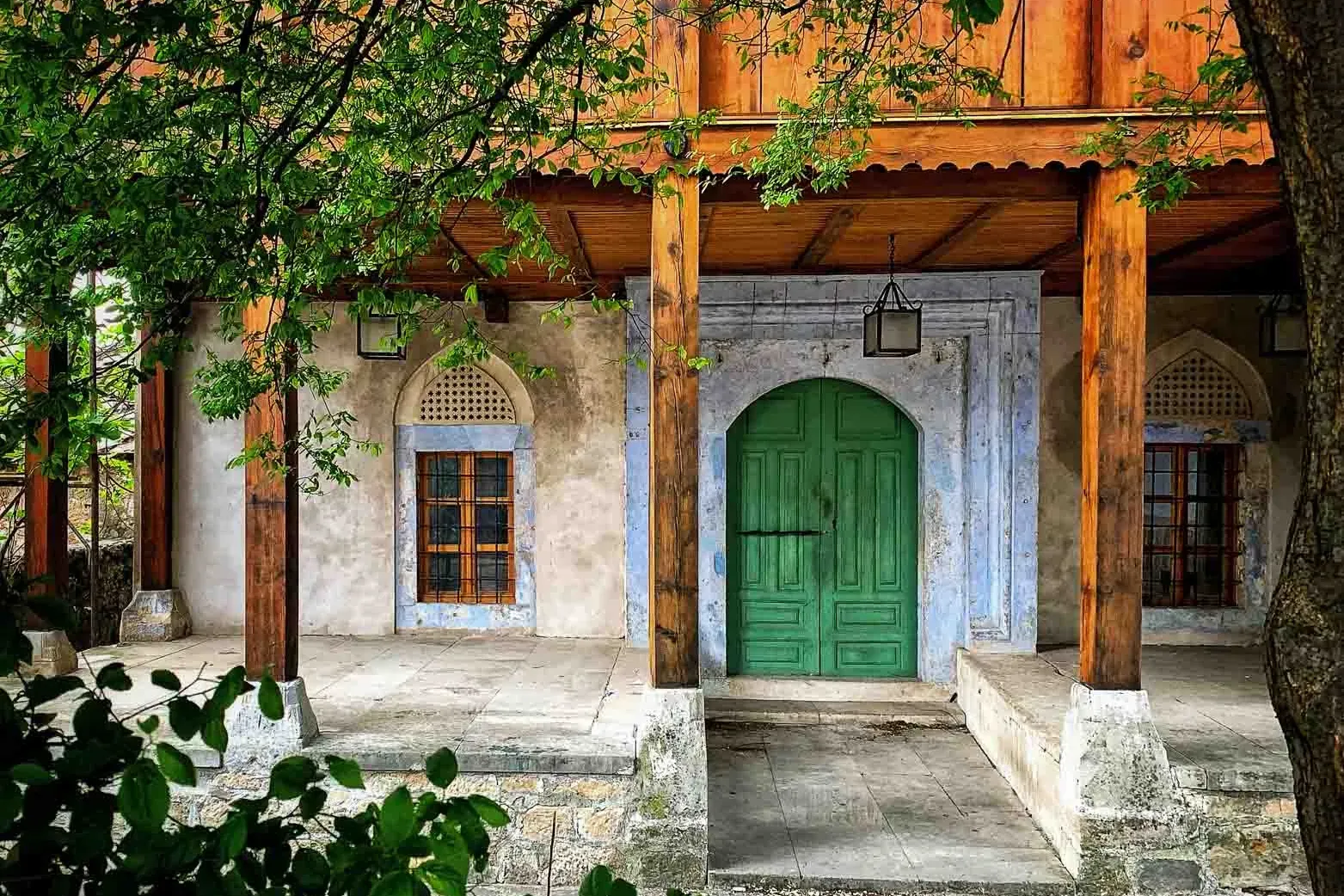 Traditional building in Mostar with a green arched wooden door, two windows, and a wooden porch supported by pillars, fram...