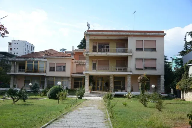 Multi-story building with light facade, closed shutters, and balconies, fronted by a paved path and green garden.
