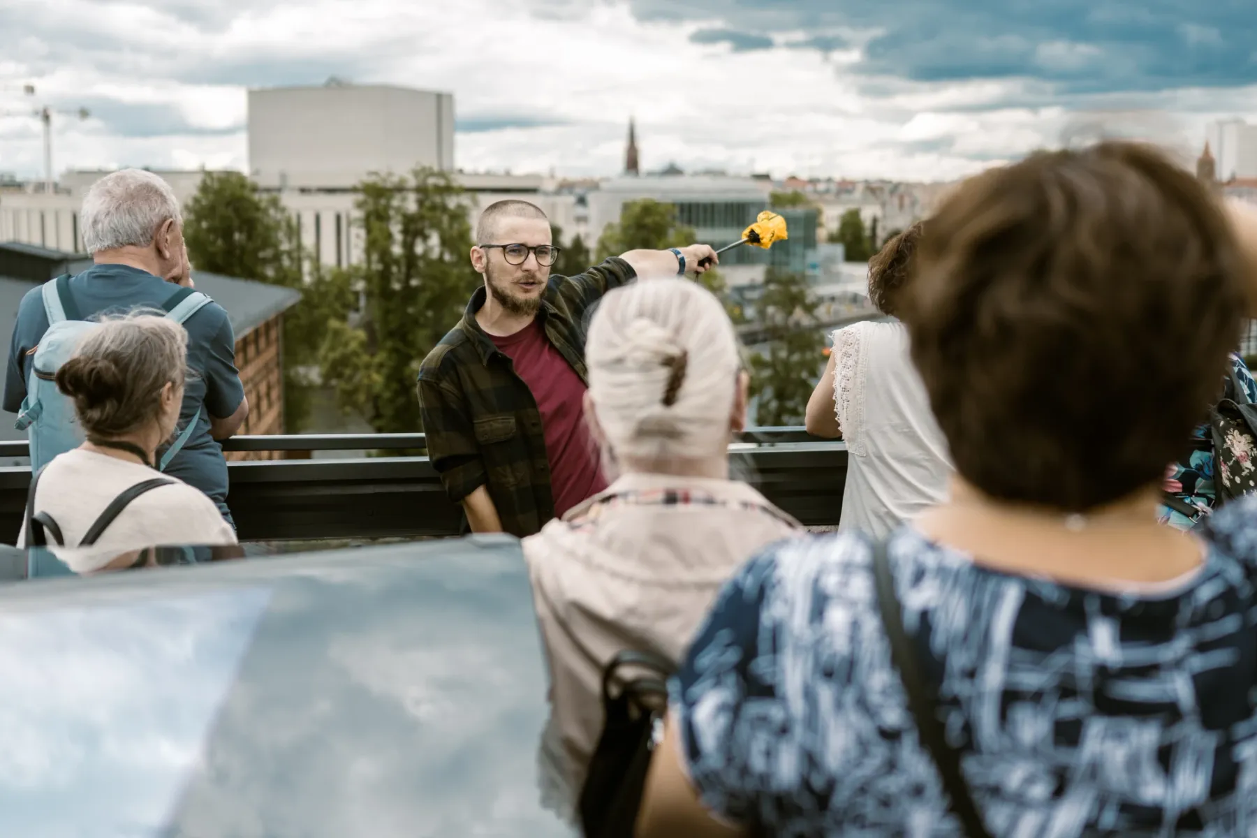 A tour guide with glasses points across the Bydgoszcz city skyline while explaining something to a small group of tourists.