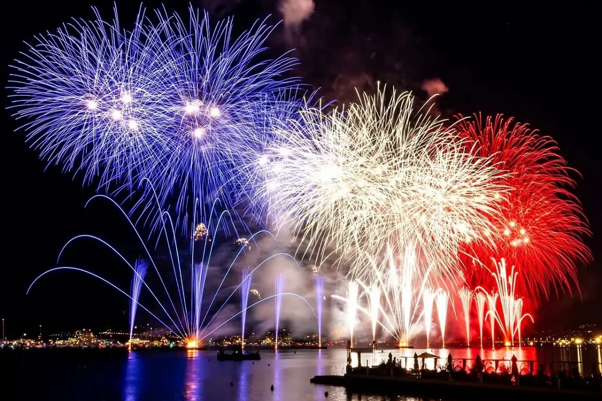Blue, white, and red fireworks explode over a bay at night. A crowd watches from the shore; city lights reflect on the water.