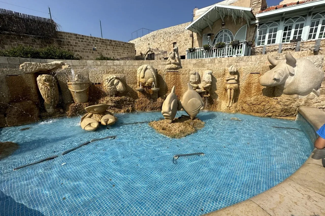 A large circular fountain with blue tiled water and a stone wall featuring various carved figures, including animals and h...