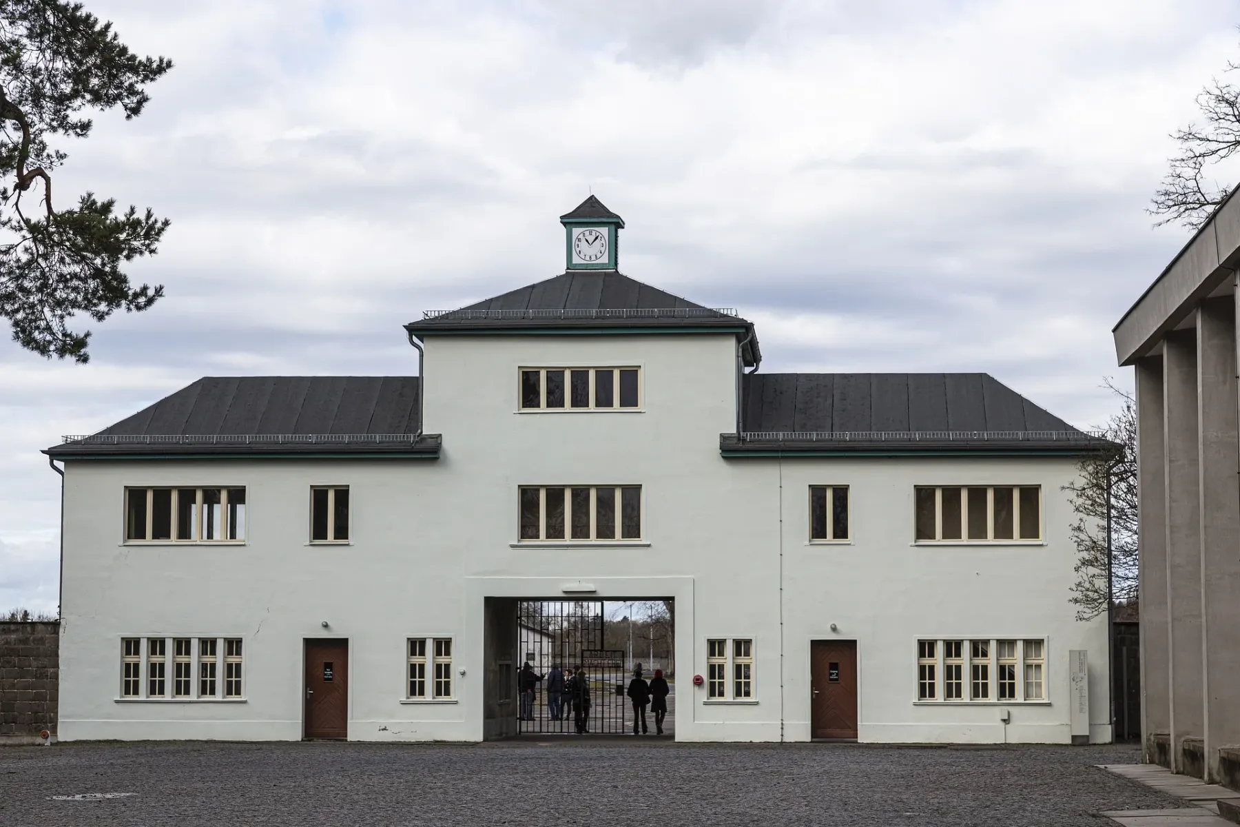 White two-story building with a clock tower and a large iron gate. People stand near the entrance on cobblestones under a ...