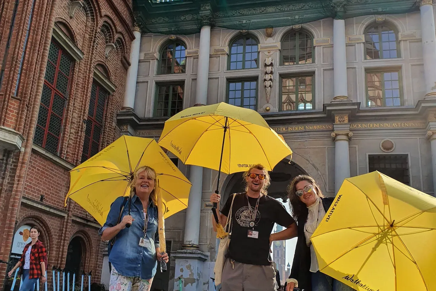Three smiling Walkative guides hold yellow umbrellas on a cobblestone street in front of Gdańsk's historic Green Gate buil...