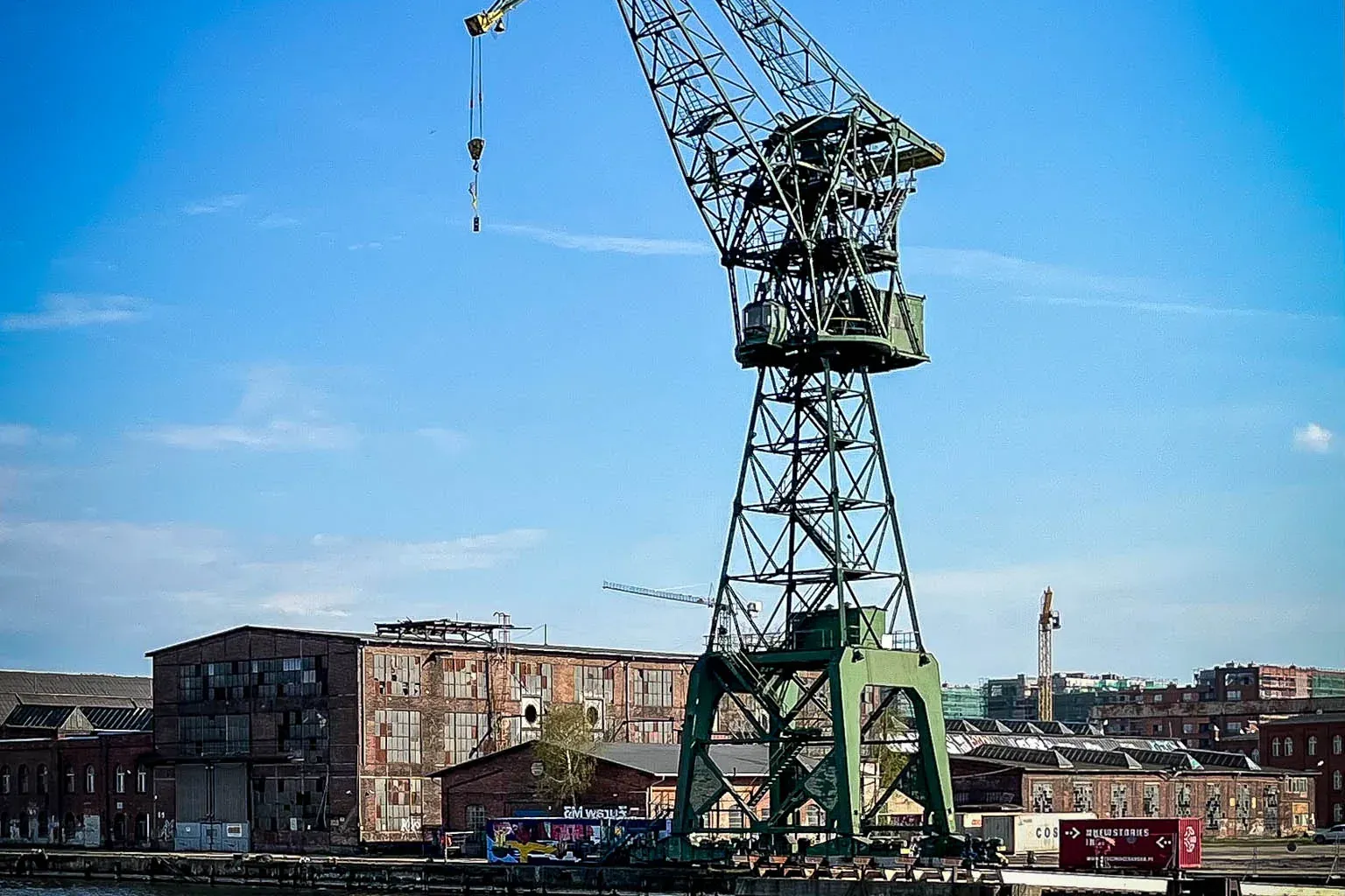 Large green gantry crane standing over the Motława River, with historic shipyard buildings under a clear blue sky in Gdańsk.