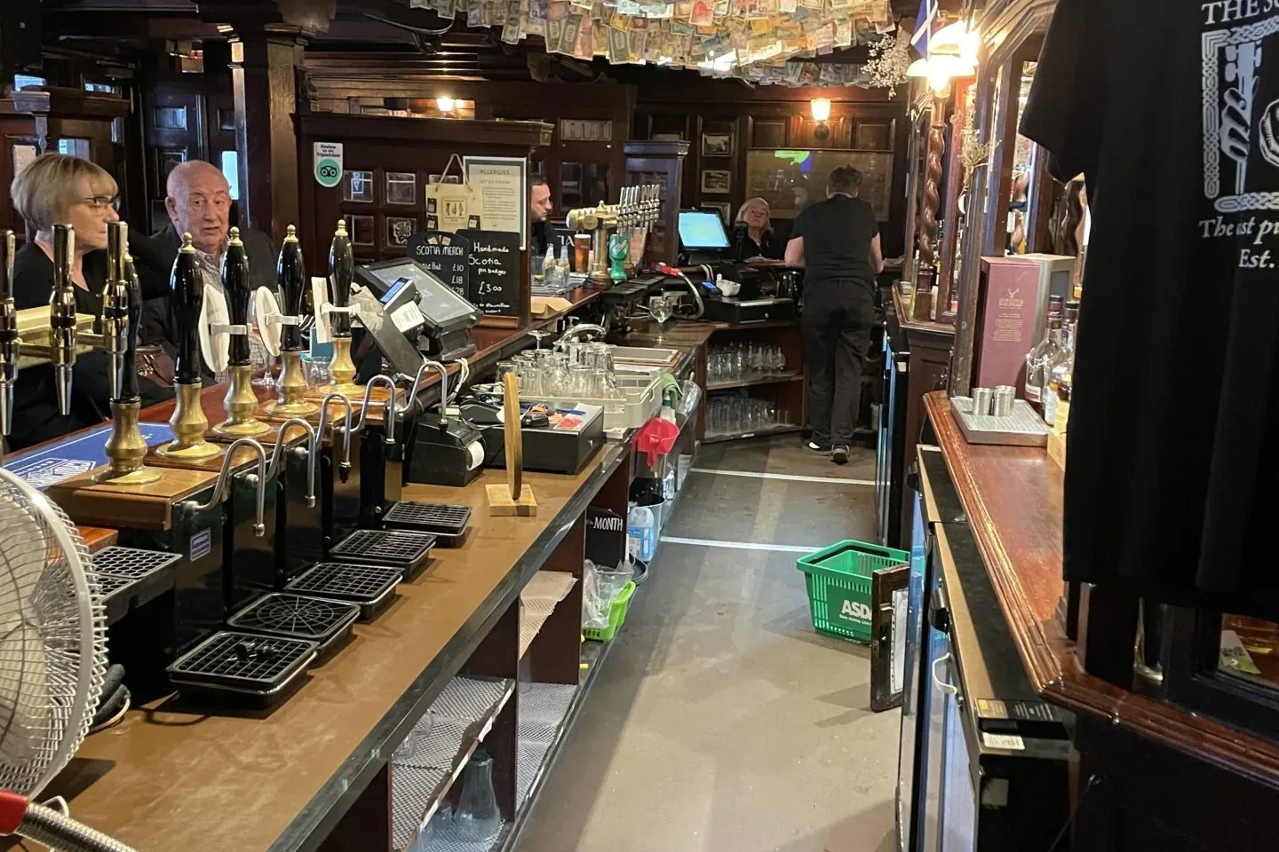 Interior of a traditional Glasgow pub with a long wooden bar, many beer taps, and banknotes from various countries strung ...