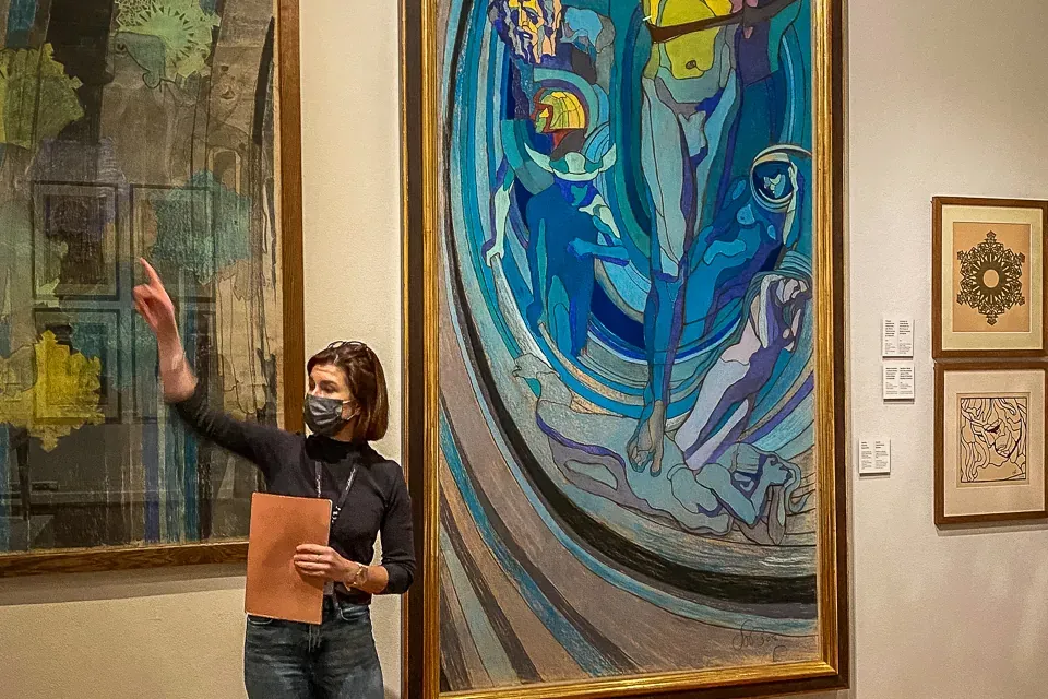 A tour guide wearing a face mask stands in an art gallery, pointing towards a large colourful painting on the wall.