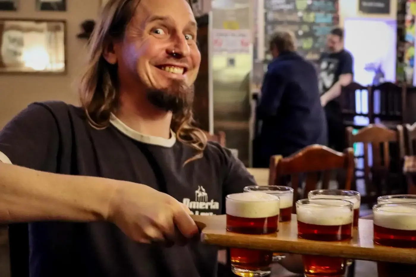 Man with long hair and beard holding a beer flight with five small glasses of amber beer in a pub.
