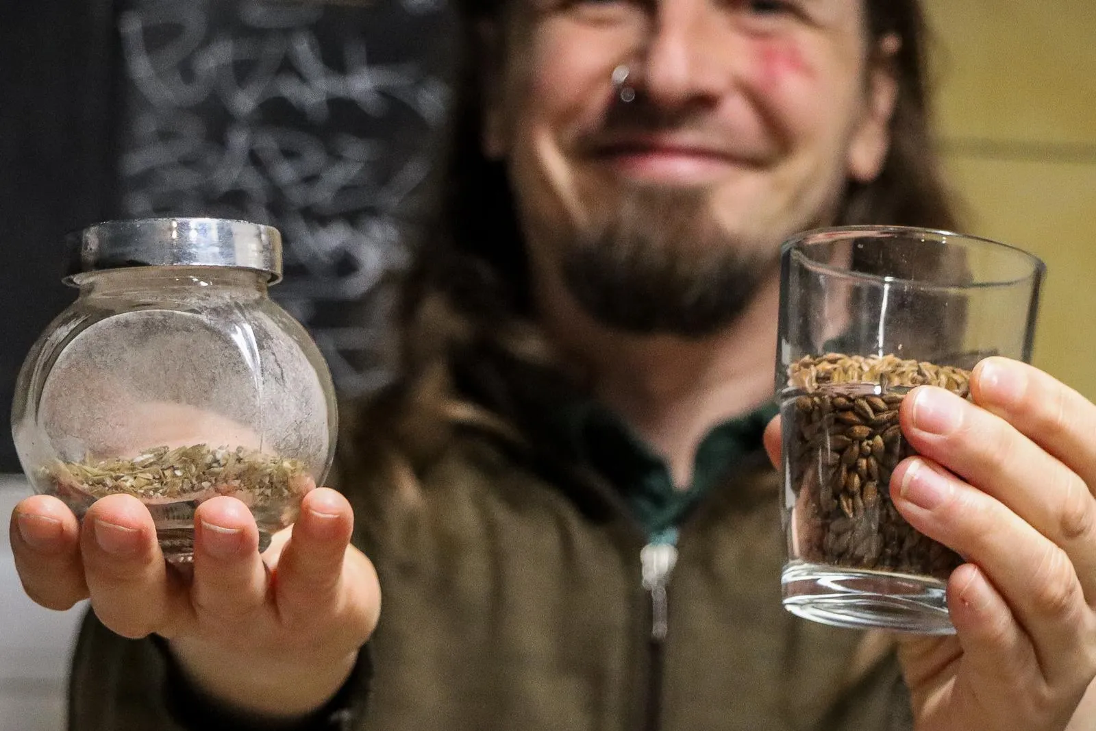 A man with long hair holds up a glass of malted barley and a small jar of dried hops toward the camera.