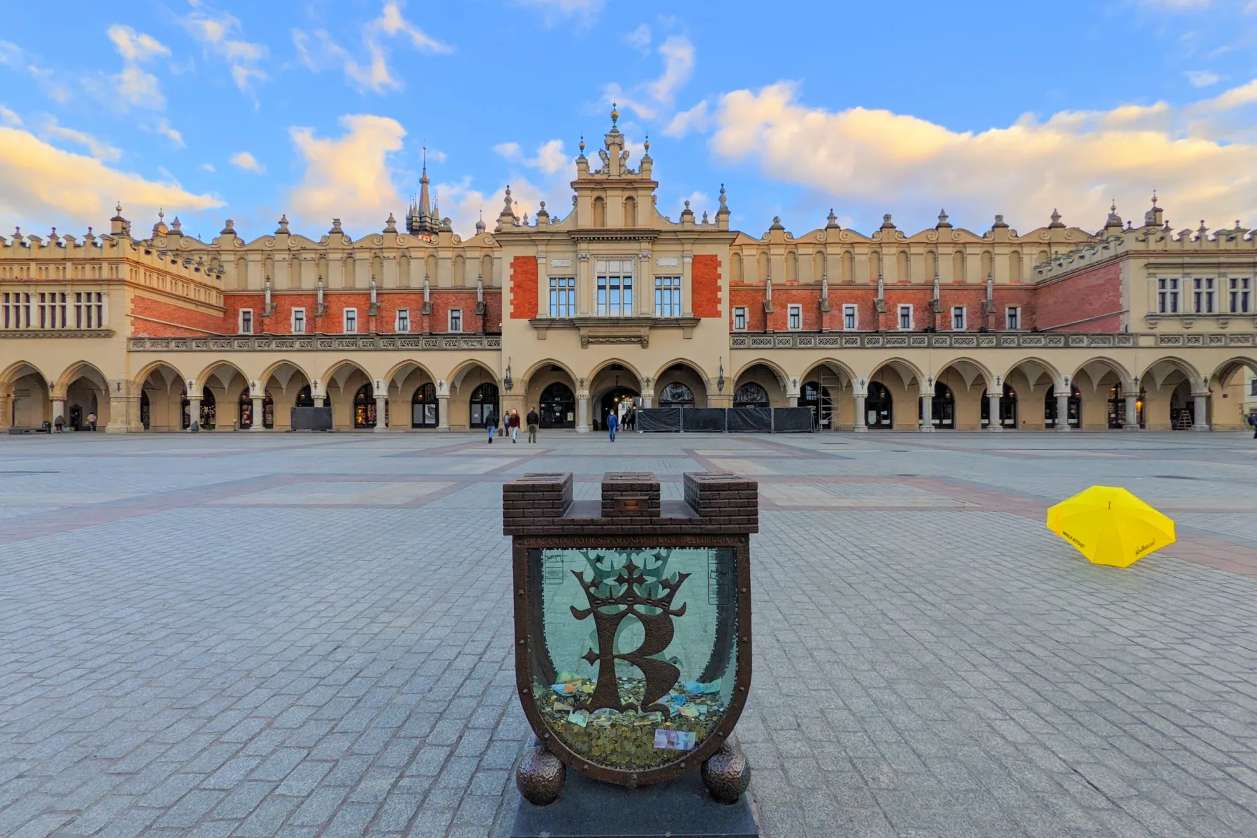 The Cloth Hall in Krakow's Main Market Square at dusk, with a donation box and a yellow umbrella in the foreground.
