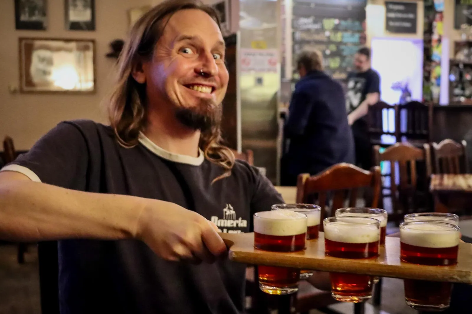 A smiling man with long hair and a nose ring holds a wooden flight with six small glasses of beer in a pub.
