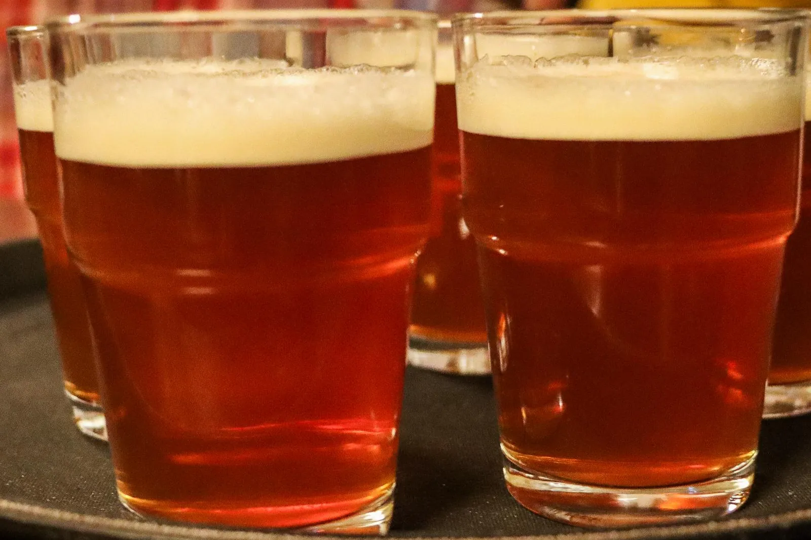 Several glasses of amber-colored beer with foam heads are arranged on a dark serving tray in a pub.